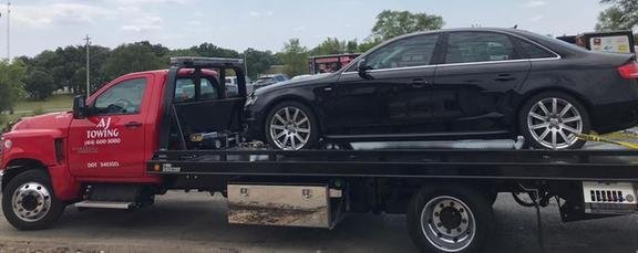 AJ Towing red flatbed tow truck with a black sedan loaded and secured on the bed.
