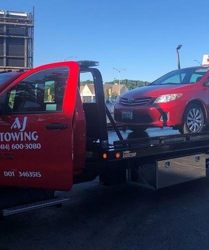 Close view of an AJ Towing flatbed door decal beside a red sedan loaded on the truck bed.